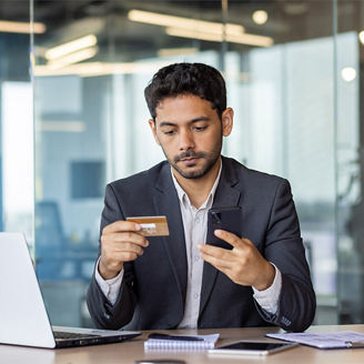 A man is sitting at a desk, holding a credit card and a mobile phone in his hands.