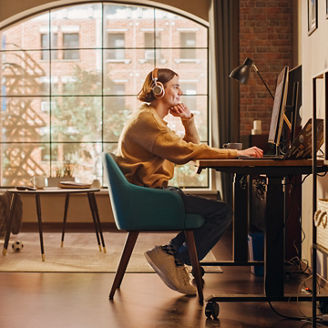 Young Handsome Man Working from Home on Desktop Computer in Sunny Stylish Loft Apartment. Creative Designer Wearing Cozy Yellow Sweater and Headphones. Urban City View from Big Window.