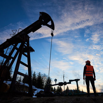 Horizontal snapshot of an oilman standing with his back to the camera, holding a pipe wrench and looking at the oil pump jack, beautiful silhouettes against blue sky, concept of oil and gas industry