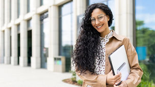 Portrait of young beautiful successful business woman outside office building, successful Latin American woman smiling and looking at camera, standing with laptop in hands and documents.