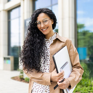 Portrait of young beautiful successful business woman outside office building, successful Latin American woman smiling and looking at camera, standing with laptop in hands and documents.