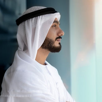 Side profile of a person wearing a white traditional head covering and robe, looking out through a window in a modern indoor setting