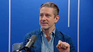 Person speaking into a microphone in a studio, gesturing while seated against a blue background with vertical light accents