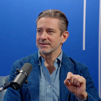 Person speaking into a microphone in a studio, gesturing while seated against a blue background with vertical light accents