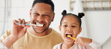 Adult and child brushing their teeth together with toothbrushes in a bright bathroom