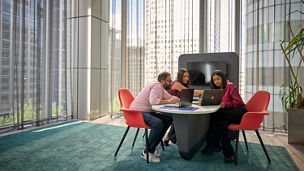 Three people gathered around a table in an office meeting area, working on laptop computers.