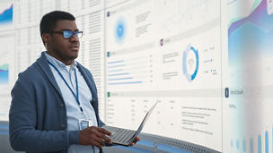Person standing with a laptop, reviewing dashboards displayed across a large screen wall