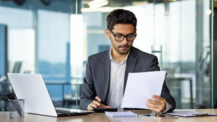 Serious concentrated hispanic businessman financier on paperwork inside office, man in business suit at workplace reviewing and reading papers, contracts and accounts reports