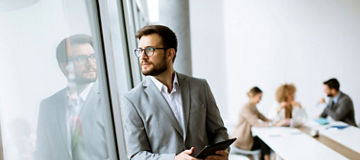Person stood looking out of an office window with people sat around a table in the background