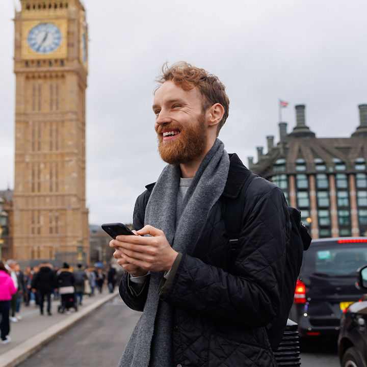 A person using a smartphone on Westminster Bridge in London with the Palace of Westminster and the Elizabeth Tower visible in the background.