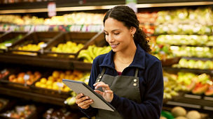 A person wearing an apron uses a tablet in the produce section of a grocery store. 