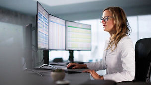 Person seated at a desk using multiple monitors displaying structured data tables
