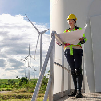 woman in a yellow helmet and safety vest is standing on a ledge looking at a piece of paper. She is wearing a reflective vest and is looking at the paper with a serious expression