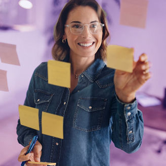 Woman wearing a denim jacket stands in an office with a glass wall that has sticky notes. Business woman engaging in a brainstorming session, showcasing her creativity and professional expertise.; Shutterstock ID 2495573727; purchase_order: marketing advisory assurance; job: Anne; client: AI campagne