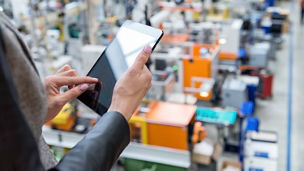 Horizontal color image of businesswoman - unrecognizable person - working with digital tablet in large futuristic factory. Woman standing on top of a balcony, holding touchpad and checking inventory of a manufacturing company on touchscreen tablet. Focus on businesswoman's hands holding black tablet, futuristic machines in background.