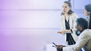 Three people gathered around a meeting table, with one person standing and gesturing while two others sit nearby, one holding a smartphone