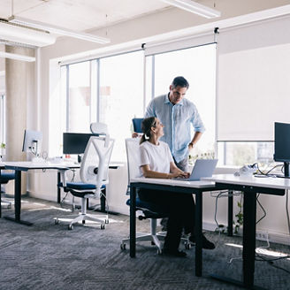 A woman is seated at her desk, working on a computer, while a standing colleague is behind her, assisting with the task