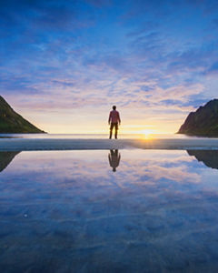 Reflected landscape of person standing between mountains looking at sunset over ocean