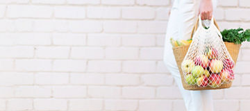 Eine Person in weißer Hose und weißen Schuhen trägt eine Einkaufstasche aus Netzmaterial, die mit verschiedenen Obstsorten und Gemüse gefüllt ist. Die Person steht vor einer weißen Ziegelwand.