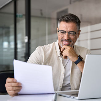 Smiling Latin or Indian male business man accountant analyst holding documents, work at laptop computer doing online trade market tech research. Focused Hispanic businessman with paperwork in office.