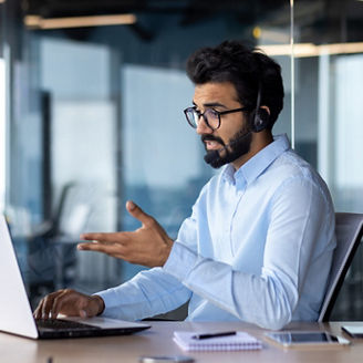 A young Indian businessman in a headset works in the office and conducts an online business meeting from a laptop, talks on a video call with clients, partners.