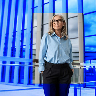 Mulher em ambiente empresarial, de pé num escritório moderno com fachadas envidraçadas azuladas.