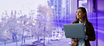 Happy businesswoman using laptop while looking through window in office.