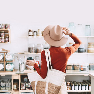 Minimalist vegan style girl with wicker bag and reusable glass coffee cup on background of interior of zero waste shop. Woman doing shopping without plastic packaging in plastic free grocery store.