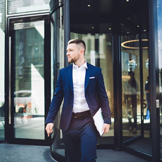 Elegant young male office worker in formal wear with clipboard leaving contemporary business centre building thoughtfully looking away in sunlight