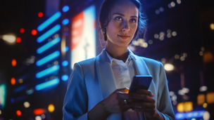 Beautiful Young Woman Using Smartphone Standing on the Night City Street Full of Neon Light. Portrait of Gorgeous Smiling Female Using Mobile Phone.