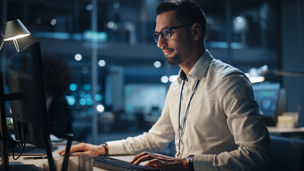 In Big Corporate Office at Night: Portrait of Confident Manager in White Shirt Using Computer, Businesspeople and Experts Working Around Him, Analysing Statistics, Commerce Data, Marketing Plans.