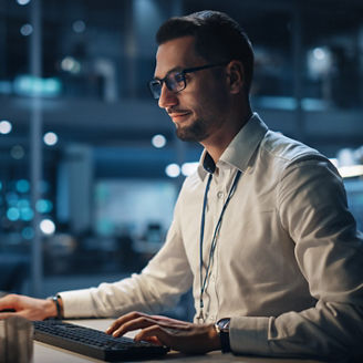 In Big Corporate Office at Night: Portrait of Confident Manager in White Shirt Using Computer, Businesspeople and Experts Working Around Him, Analysing Statistics, Commerce Data, Marketing Plans.