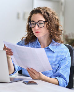 Young busy business woman manager, lawyer or company employee holding accounting bookkeeping documents checking financial data or marketing report working in office with laptop. Paperwork management