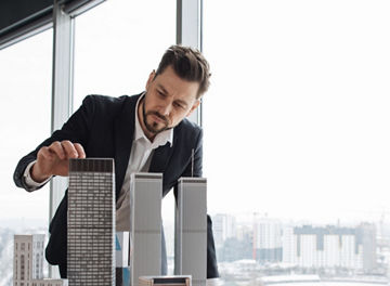 Confident businessman real estate agents in black stylish formal suit standing while touching 3d model skyscraper of city architecture design at office with panoramic city view.