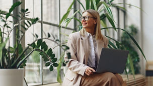Smiling woman programmer in eyeglasses working on laptop while sitting in coworking near window