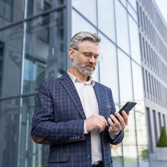 Portrait of a senior gray-haired male doctor standing in a suit outside the office and using a mobile phone, reading the news, typing a message, waiting for an appointment.; Shutterstock ID 2295143863; purchase_order: 