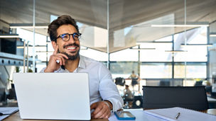 Happy Latin business man working at laptop in office looking away. Happy young businessman professional using computer sitting at desk thinking of corporate technology ai solutions. Copy space