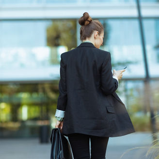 Seen from behind middle aged business woman near office building in black jacket with smartphone and briefcase walking.