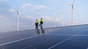 Engineers work walking on factory roof inspect survey and check solar cell panel by hold boxed with turbine wind mill field background is ecology energy alternative power factory concept.