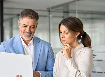 Busy professional team of two middle aged business people company executives standing in office using laptop computer tech device discussing corporate financial project results working together.