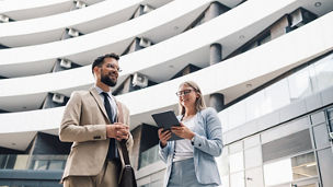 Two young professionals standing in front of a modern office building, engaging in conversation while looking at a tablet
