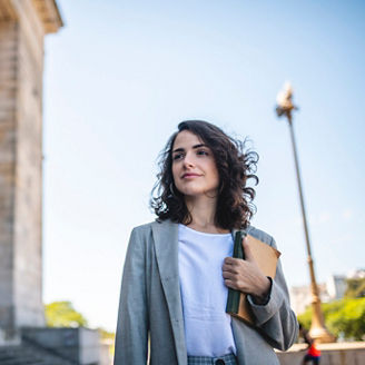 Low angle view of female law student in late 20s standing with book and pensive expression at entrance to Neo-Classical university building.