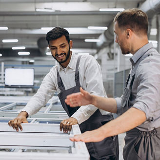 Two male international workers in overalls work together at a machine for the production of aluminum PVC windows and doors