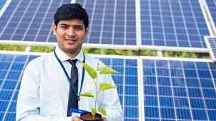 banker holding tree plant in hand in front of solar panel by looking at camera - concept of environmental growth, sustainable lifestyle and renewable energy.