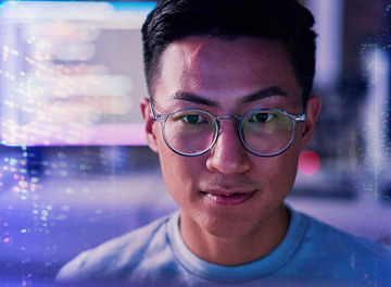 Close-up of a man wearing round glasses, facing forward, with blurred blue and purple digital light patterns and interface elements in the background