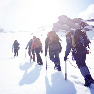 Das Bild zeigt eine Gruppe von Personen, die in einer verschneiten Berglandschaft wandern. Sie tragen Winterkleidung und Rucksäcke und verwenden Wanderstöcke.
