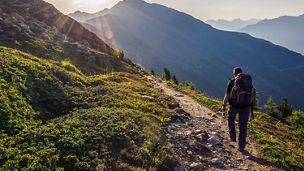 Das Bild zeigt eine Person, die auf einem Wanderweg in einer bergigen, grünen Landschaft geht. Sie trägt einen Rucksack und ist von Vegetation umgeben. Die Sonne scheint tief am Himmel und beleuchtet die Szene.