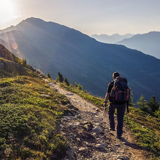 Das Bild zeigt eine Person, die auf einem Wanderweg in einer bergigen, grünen Landschaft geht. Sie trägt einen Rucksack und ist von Vegetation umgeben. Die Sonne scheint tief am Himmel und beleuchtet die Szene.