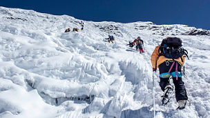 Mehrere Personen klettern eine schneebedeckte und vereiste Bergwand hinauf. Sie tragen Kletterausrüstung, einschließlich Steigeisen und Seilen, während der Himmel klar und blau ist.