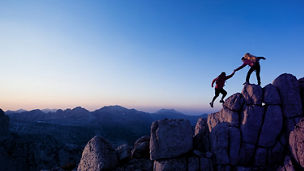 Zwei Personen klettern auf einem Felsen bei Sonnenuntergang, wobei eine Person der anderen beim Aufstieg hilft, während im Hintergrund eine Berglandschaft sichtbar ist.